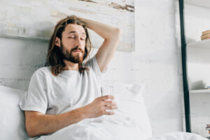young bearded man with long hair having hangover and taking pill with glass of water in bedroom at home